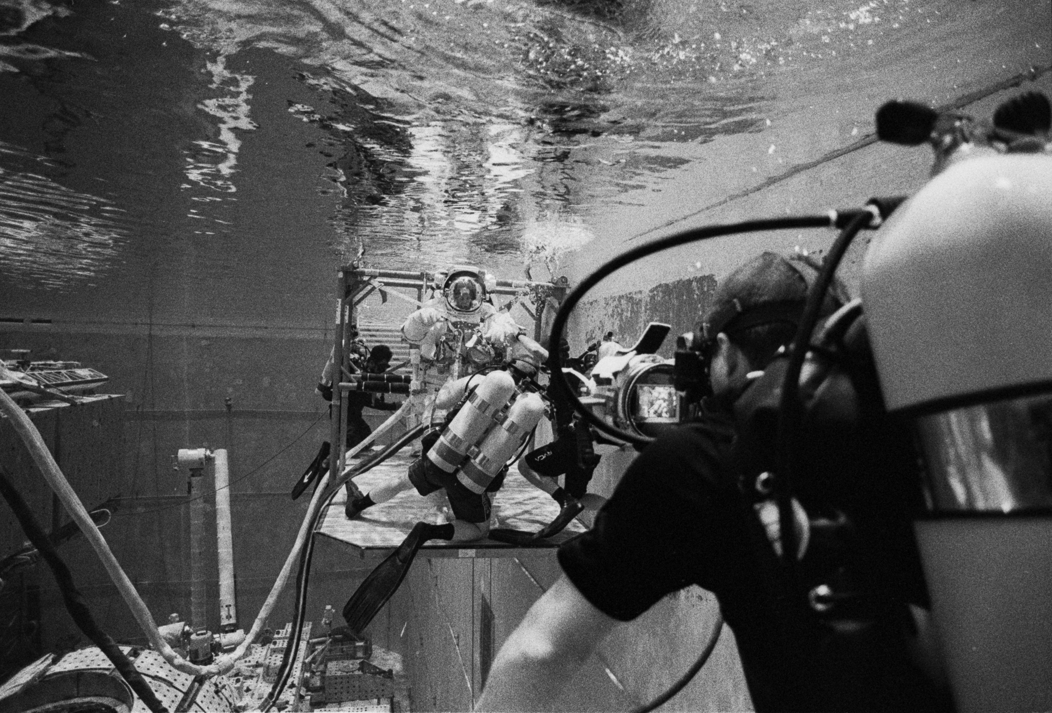 NASA crew members are lowered into the pool via a jib crane and secured into a donning stand. The divers await commands given by the test director before beginning egress of the donning stand. Photo by Moe Lauchert.